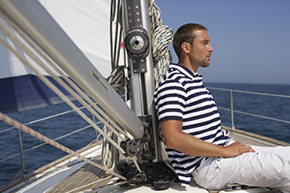 Ein Mann in einem blau-weiß gestreiftem Poloshirt sitzt auf einer Segelyacht und lehnt sich an den Mast.