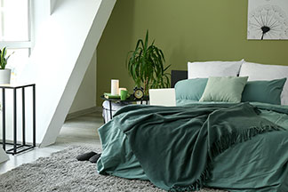 Schlafzimmer Deko farblich abstimmen: Ein modernes Schlafzimmer mit Tagesdecken und Kopfkissen in verschiedenen Grüntönen, Pflanzen und einem weißen Bild mit Pusteblume an der Wand über dem Bett.