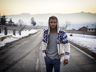 Ein junger Mann, der eine weiße Strickjacke mit Norwegermuster und Kapuze trägt, läuft bei Sonnenaufgang auf einer winterlichen Straße durch eine nebelverhangene Landschaft.