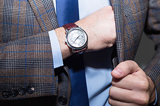 Herrenuhren als Hingucker: Nahaufnahme einer analogen Uhr am Arm eines Mannes, der ein hellblaues Hemd und ein kariertes Sakko trägt.