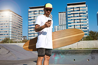 Ein Skater mit Sonnenbrille und Cap steht in einem Oversized-Shirt und seinem Longboard in der Hand in einem Skatepark und schaut auf sein Handy.