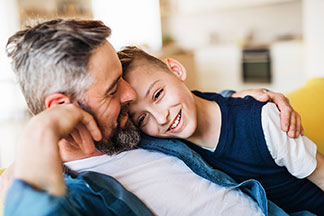 Qualität bei Tchibo für die Familie: Ein Vater in einem offenen Jeanshemd sitzt mit seinem Sohn in den Arm gekuschelt auf dem Sofa.