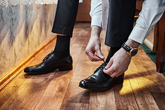 Nahaufnahme der Beine eines Mannes, der eine schwarze Anzughose, schwarze Lederschuhe und schwarze Herrensocken trägt, auf seinem Bett sitzt und sich gerade die Schuhe bindet.