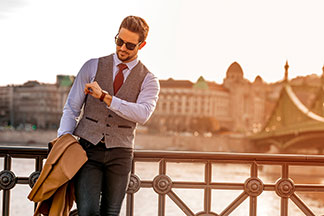 Ein elegant gekleideter Herr mit Sonnenbrille schaut auf seine Herrenuhr. Dabei steht er vor einem Fluss und lehnt an einem Geländer.