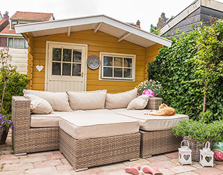 Gartenhaus als Wohlfühloase: Vor einem Gartenhaus aus Holz steht eine große Couch aus Rattan mit weißen Sitzauflagen und Kissen.