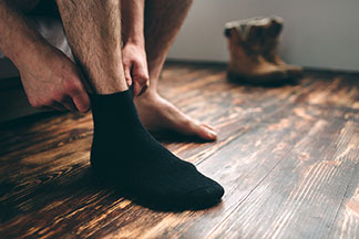 Nahaufnahme der Beine eines Mannes, der auf seinem Bett sitzt und sich seine Strümpfe anzieht. Ein Fuß trägt schon einen schwarzen Socken, der zweite ist noch barfuß.