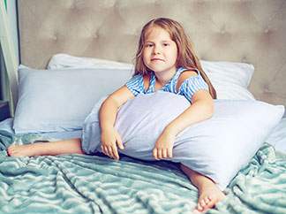 Klassiker unter den Kopfkissen: Ein Mädchen sitzt mit einem schmalen, rechteckigen Kissen im Arm auf einem Bett.