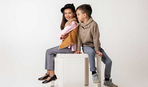 Zwei stylische Kinder sitzen auf einem Tisch. Der Junge schaut das Mädchen verschmitzt an.