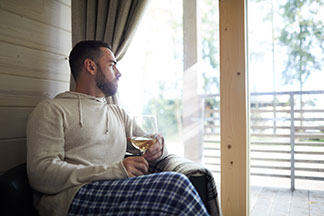 Ein junger Mann, der Homewear bestehend aus einer karierten Hose und einem hellen Pullover mit Kapuze trägt, sitz am Fenster und trinkt ein Glas Wein.