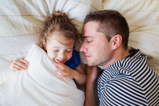 Keine Sorgen dank Tchibo-Qualität: Ein Vater in einem geringelten Sweater liegt zusammen mit seiner Tochter im elterlichen Bett und schläft.