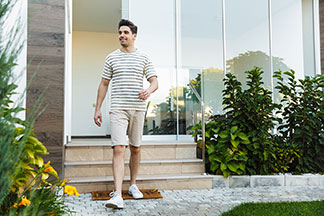Ein junger Mann in einem schwarz-weiß gestreiften Oberteil und beigen Chino-Shorts verlässt beschwingt sein Haus.