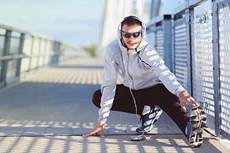 Sportjacken für alle Situationen: Ein junger Mann in einer weißen Laufjacke macht mit Kopfhörern Stretch-Übungen auf einer Brücke.