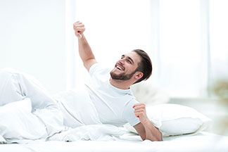 Ein junger Mann, der ein weißes T-Shirt trägt, liegt am Morgen noch halb unter seiner weißen Decke und streckt sich zufrieden in seinem Bett.