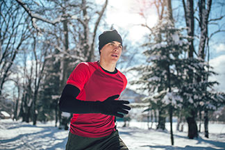 Ein junger Mann, der ein rotes Sportshirt und darunter ein langärmliges schwarzes Unterhemd sowie eine schwarze Mütze trägt, joggt im Winter im Freien.