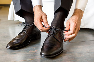 Die richtigen Herrenstrümpfe fürs Büro: Nahaufnahme eines Mannes mit dunkler Anzughose und dunkelbraunen Strümpfen beim Binden seiner dunkelbraunen Lederschuhe.