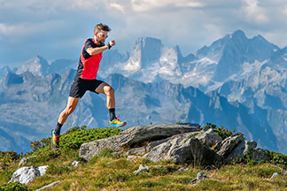 Ein junger Mann, der kurze, schwarze Sporthosen und ein rot-schwarzes Shirt trägt, joggt bei schönem Wetter durch eine Berglandschaft.