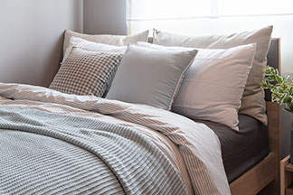 Kissenbezüge aus hochwertigen Materialien: Ein Bett mit verschieden großen Kopfkissen in hellen Erdtönen, einer gestreiften Bettdecke und einer hellblauen Tagesdecke.