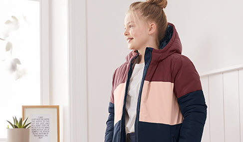 Passt perfekt: ein junges Mädchen steht im Hausflur und trägt eine rosa-rot-blau-gestreifte Jacke.