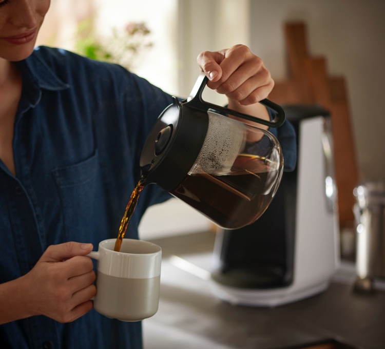 Eine Frau trinkt genüsslich ihren Kaffee aus einer Filterkaffeemaschine, die im Hintergrund zusehen ist.
