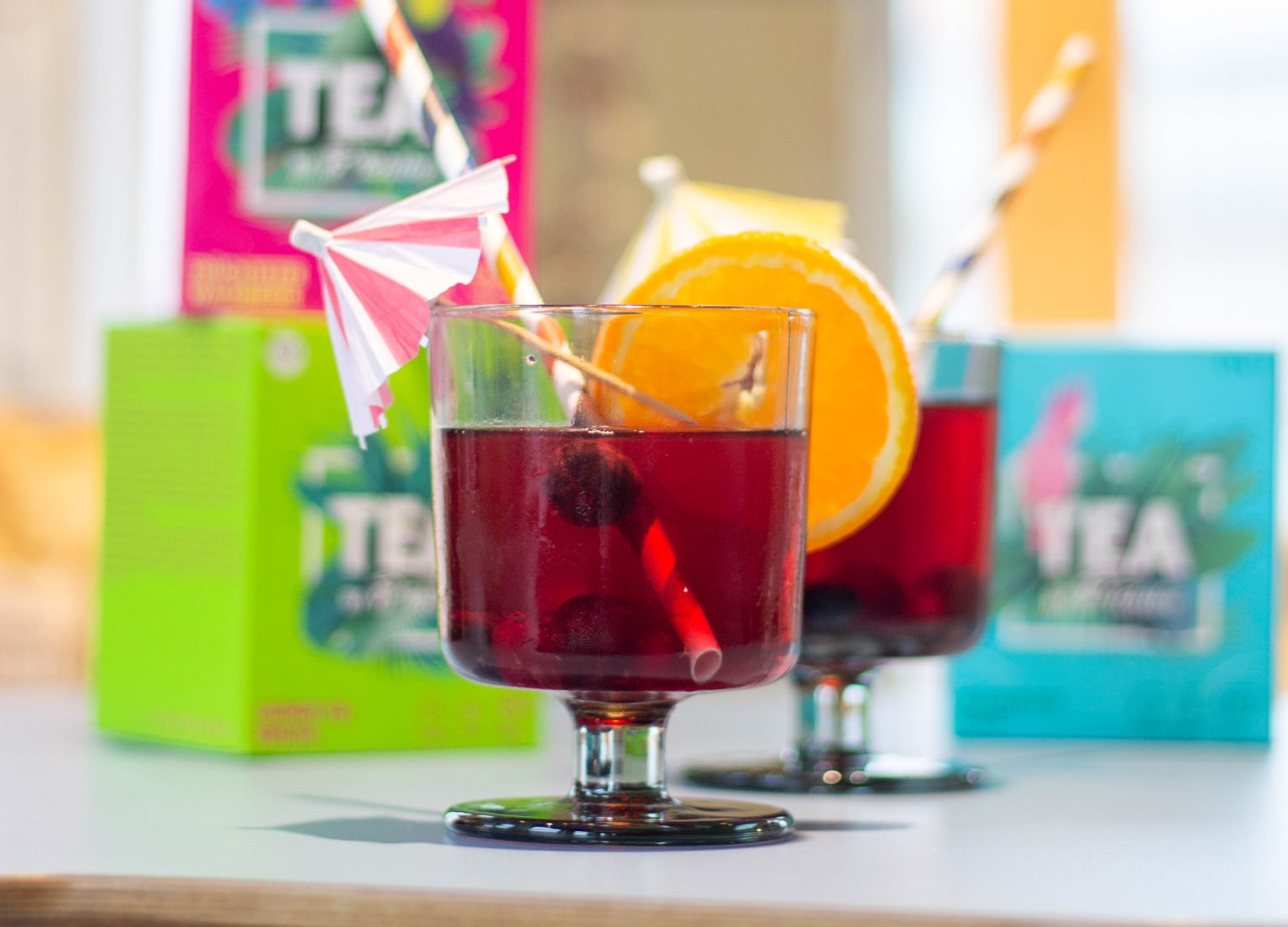 Cocktailglas mit Heidelbeeren, Orange und Strohhalm mit Tchibo-Tee im Hintergrund