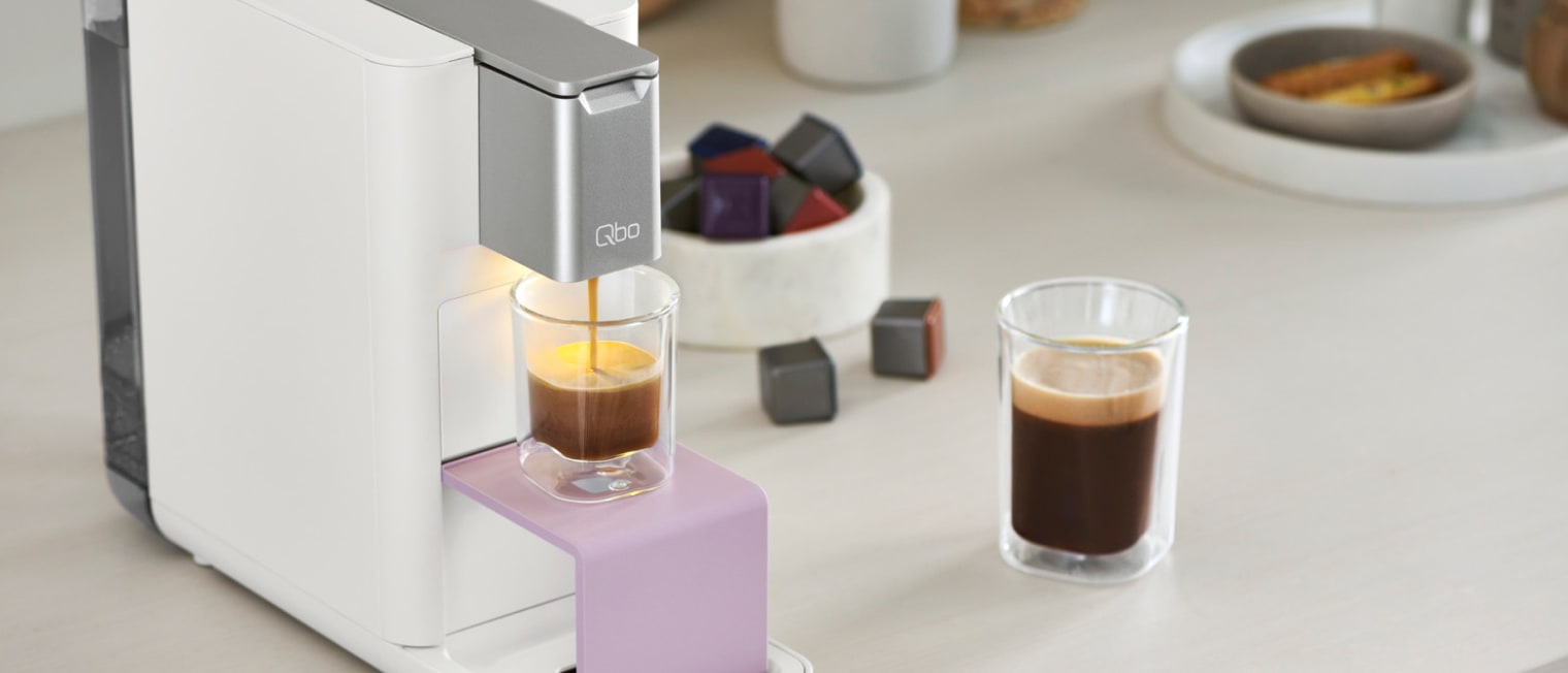 Eine Qbo Kapselmaschiene, die auch einem Tisch mit einem frisch zubereiteten Kaffee steht.','Enter picture alt'}