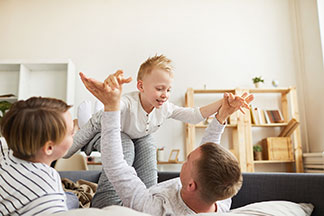 Eine junge Familie liegt in Homewear auf dem Bett und der Vater lässt den kleinen Sohn auf seinen Beinen wie ein Flugzeug fliegen.