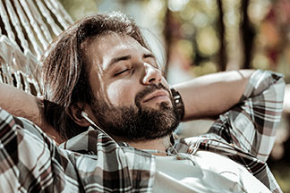 Gute Qualität bei Tchibo-Basics: Ein bärtiger Mann liegt mit weißen In-Ear-Ohrhörern und einem Holzfällerhemd in einer Hängematte und hat die Augen geschlossen.