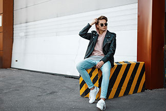 Herren Halbschuhe für den Sommer: Ein junger Mann in Jeans, Kapuzenpullover und weißen Sneakers sitzt lässig auf einem schwarz-gelb gestreiften Betonblock vor einer Garage.