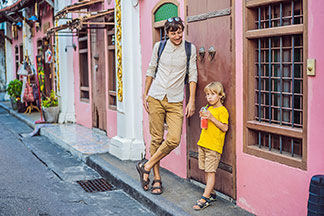 Herrenshorts in allen Varianten: Ein Vater in 3/4-Shorts steht mit seinem Sohn, der eine Shorts bis zum Knie trägt, an eine Hauswand gelehnt.