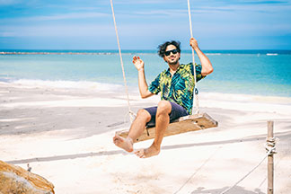 Freizeitlook: Ein bärtiger Mann in einem kurzärmeligen Hawaiihemd sitzt auf einer Schaukel am Strand. Im Hintergrund ist das Meer zu sehen.