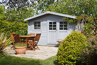 Loungemöbel im Gartenhaus lagern: In einem Garten mit hohen grünen Hecken steht ein graues Gartenhaus, vor dem ein runder Holztisch mit vier Holzstühlen steht.