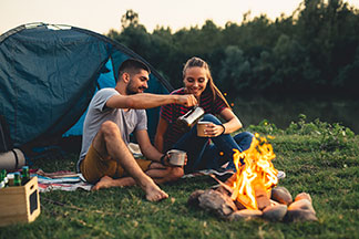 Die richtige Ausrüstung: Ein junges Paar in Outdoorbekleidung sitzt gut gelaunt vor seinem Zelt am Feuer und trinkt Kaffee.
