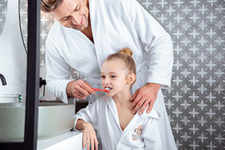 Ein Vater und Tochter, die beide jeweils einen weißen Bademantel tragen, stehen im Badezimmer und der Vater putzt der Tochter die Zähne