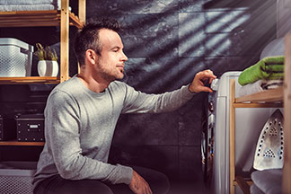 Sporthose richtig waschen: Vor einer Waschmaschine kniet ein Mann und stellt das Waschprogramm ein..