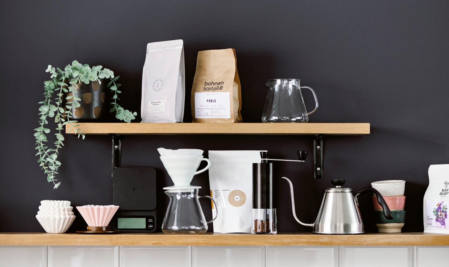 Zwei Regalbretter sind an einer dunkelgrauen Wand befestigt und es stehen drei Packungen Specialty Coffee darauf sowie verschiedene Kaffeezubereiter und eine Pflanze.