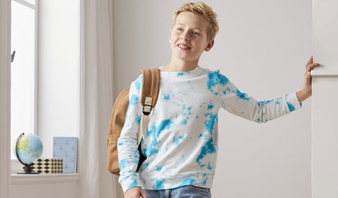 Ein Junge in einem blau-weiß-melierten Pullover wartet mit seinem Rucksack im Flur.