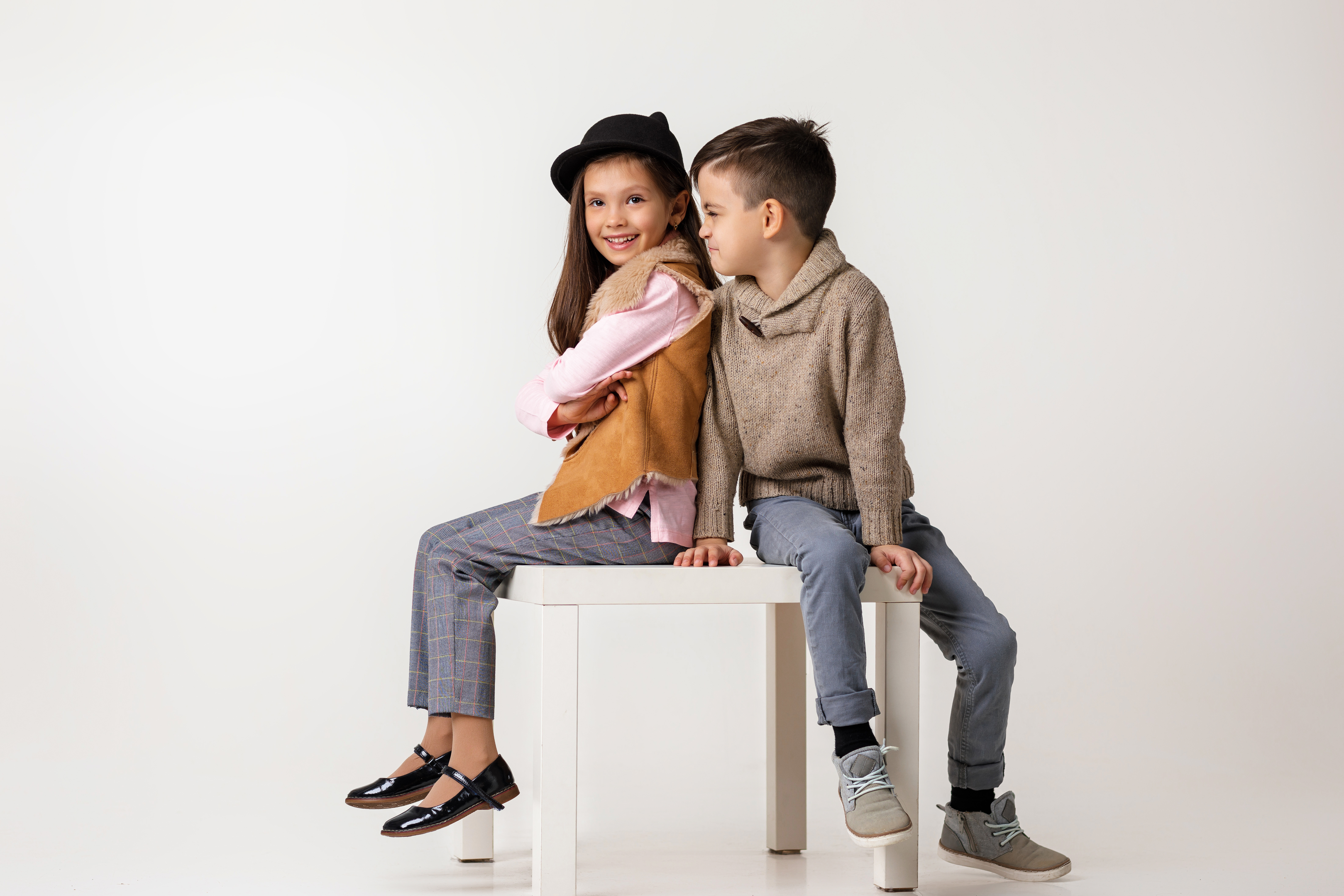 Zwei stylische Kinder sitzen auf einem Tisch. Der Junge schaut das Mädchen verschmitzt an.