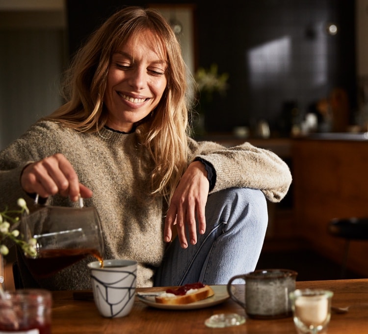 Eine Frau sitzt entspannt an einem Tisch und schenkt sich Kaffee in eine Tasse ein.