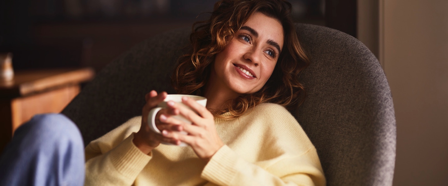 Stimmungsvolles Bild einer Frau, die lachend auf einem Sessel liegt und den Moment mit ihrem Kaffee genießt.