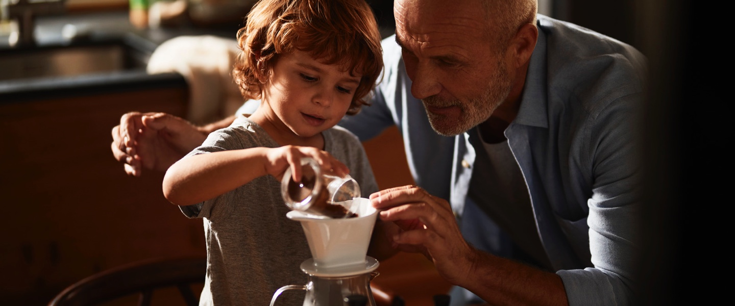 Stimmungsvolles Bild zwischen Mann und Kind, wobei das Kind gemahlenen Kaffee in einen Filter schüttet.