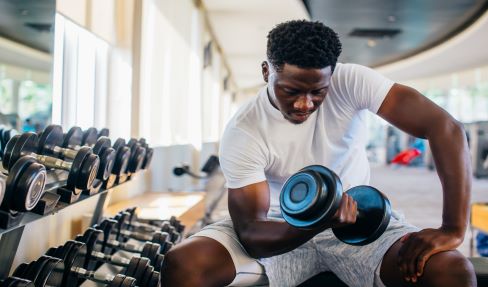 Nach dem Sport die richtige Pflege: Ein junger Mann sitzt im Sportstudio und drückt in einem weißen Sportshirt Hanteln.