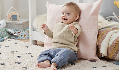 Günstige Kleidung für ihr Baby: Ein Baby sitzt in einem Shirt und einer gestreiften Hose und lehnt sich an ein Kissen.