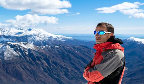 Ein Mann, der eine blaue Skijacke und eine Skibrille trägt, steht auf der Piste in einer verschneiten Berglandschaft.