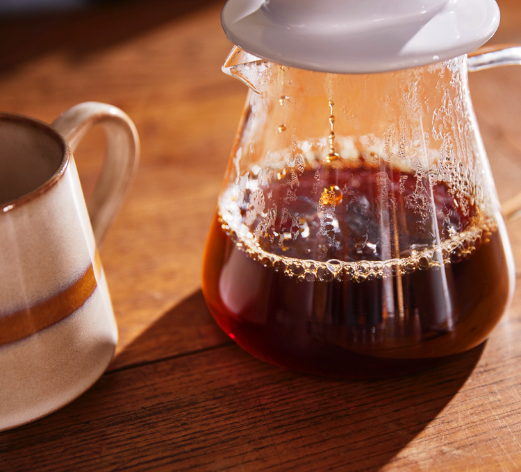 Eine Frau sitzt entspannt an einem Tisch und schenkt sich Kaffee in eine Tasse ein.