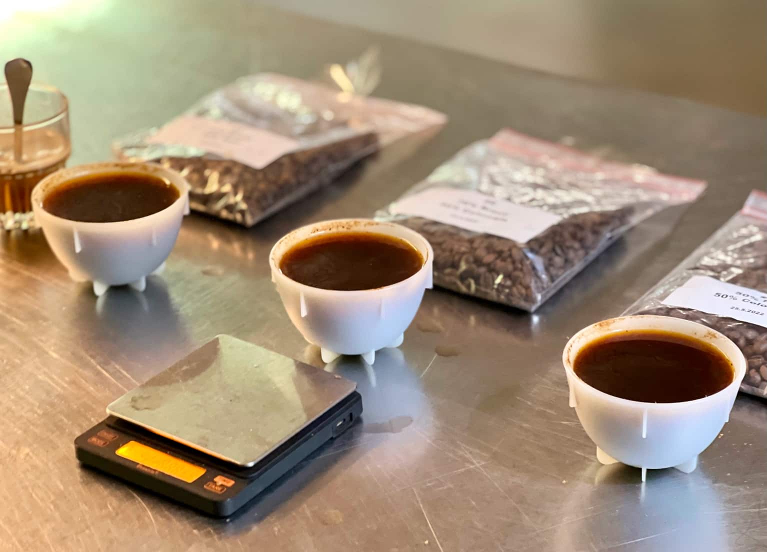 Drei Cuppingtassen sind mit Specialty Coffee gefüllt und stehen neben einer Waage und Kaffeepackungen auf einem Tisch