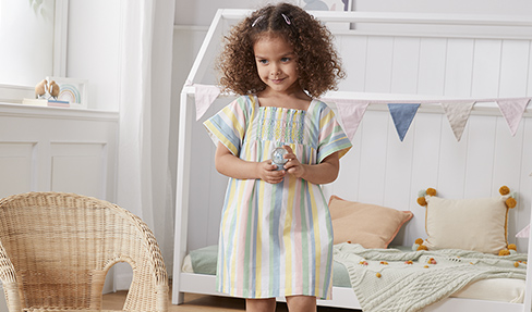 Kindersachen in allen Farben: Ein kleines Mädchen steht in ihrem gestreiften Kleid vor ihrem Bett
