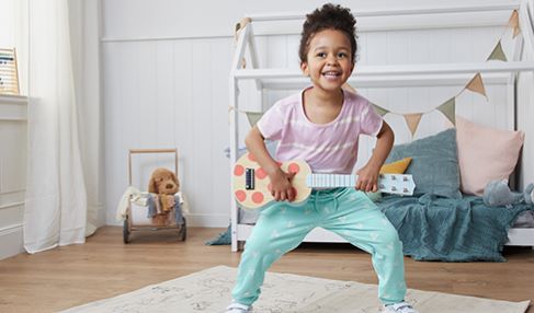 Ein kleines Mädchen spielt mit einer Spielzeuggitarre. ihr Kinderbett ist mit Fähnchen geschmückt.