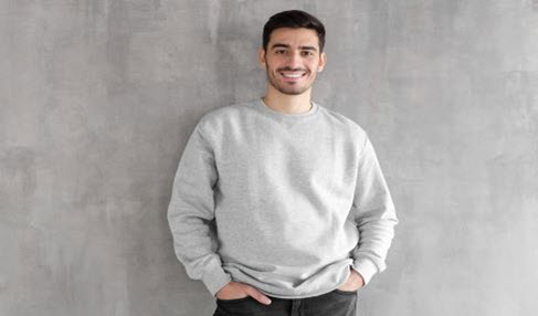 Junger Mann trägt einen hellgrauen Pullover und lehnt sich lächelnd gegen eine Wand.