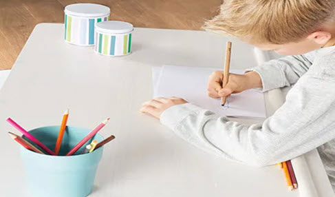 Ein kleiner junge sitzt an einem höhenverstellbaren Kinderschreibtisch und macht Hausaufgaben.
