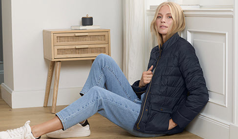 Frau steht in hellblauer Bluse und Jeans und hält sich mit der Hand an einem Regal fest.